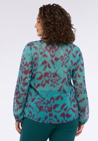 Donna con capelli ricci che indossa una blusa teal con motivi astratti viola e pantaloni teal scuri, di spalle contro uno sfondo bianco.