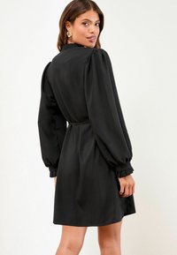 Schwarzes Kleid mit langen, puffigen Ärmeln, einer leichten Taillierung und Rüschendetails an den Manschetten. Glatter Stoff mit taillierter Passform.