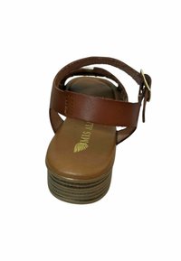 Sandalia de cuero marrón con una correa ancha y hebilla en el tobillo, con una suela lisa y un tacón marrón en capas y texturizado. Logotipo dorado en la plantilla.