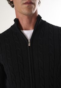 Maglione nero a zip realizzato in maglia, caratterizzato da texture a trecce, colletto alto e zip in metallo argentato. Indossato sopra una maglietta bianca.