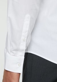 Camicetta bianca a maniche lunghe con colletto dritto, polsini con bottoni e una texture liscia; abbinata a pantaloni grigi.