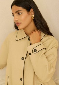 Femeie cu păr lung și închis la culoare poartă un palton bej cu nasturi cu margine neagră, ținând gulerul aproape de gât, iar o cercei aurii în formă de scoică sunt vizibili.