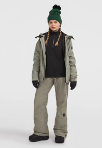 Grøn snejakke, sort lynlåsjakke, strukturerede grå bukser, sorte handsker og en grøn hue med pompon. Sorte vinterstøvler fuldender outfittet.