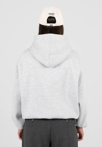 Šedá kapucová mikina s klokaní kapsou, měkká na dotek, nošena s tmavě šedými kalhotami. Béžová čepice s viditelným logem "MODERN YUPPIES".