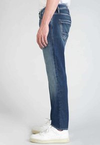 Jeans de denim en un tono azul oscuro desgastado, con un corte de pierna recta, cinco bolsillos y sutiles acentos de costura. Llevados con zapatillas blancas.
