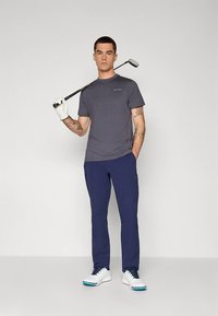 Pilka trumpomis rankovėmis golfo marškinėliai, tamsiai mėlynos kelnės ir balti golfo batai. Vyras laiko golfo lazdą; dėvi baltą pirštinę. Paprastas dizainas, minimalus prekės ženklo ženklinimas.