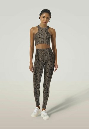 Jonge vrouw staand, gekleed in een bijpassende croptop en legging met slangenprint en witte sneakers tegen een effen achtergrond.