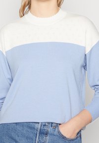 Ljusblå stickad tröja med vit scallopad rund hals och mönstrad övre del. Mjuk textur, långa ärmar, avslappnad passform.