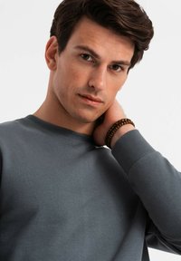 Grauer Pullover mit Rundhalsausschnitt und gerippten Bündchen. Das Modell trägt ein Holzperlenarmband am Handgelenk und fasst sich an den Hals. Neutraler Hintergrund.