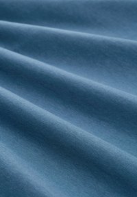 Modrá látka s hladkým texture a jemným leskom, s jemnými záhybmi a vlnami, ktoré zdôrazňujú konzistentný svetlý a tmavý gradient.