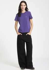 T-shirt di cotone viola con collo rotondo, abbinato a pantaloni in denim nero a larga vestibilità. I dettagli chiave includono un piccolo logo sulla t-shirt.