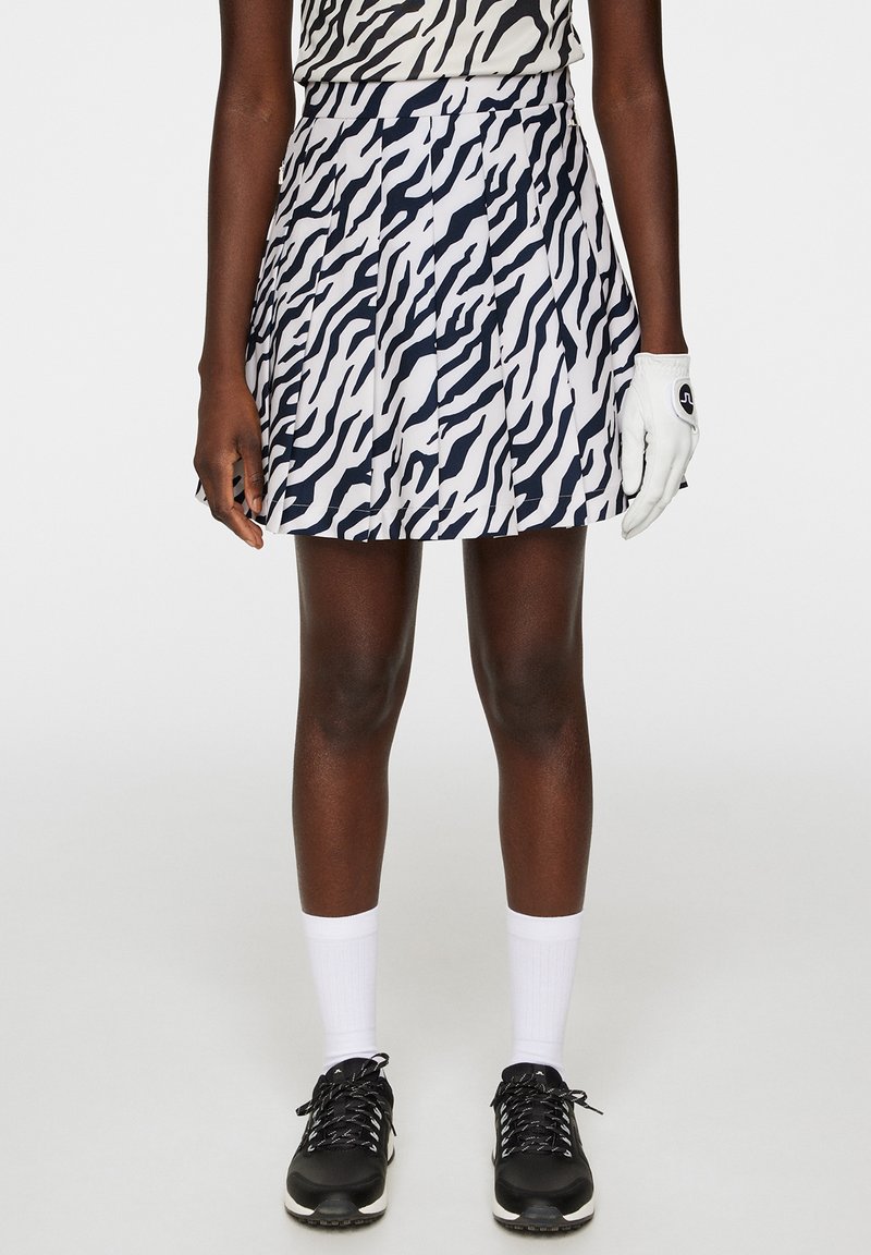 Gonna da golf plissettata bianca con stampa zebra blu navy, realizzata in tessuto leggero. Indossata con scarpe nere e calze bianche, abbinata a un guanto coordinato.