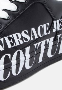 Μαύρο δερμάτινο αθλητικό παπούτσι με λευκά εκτυπωμένα γράμματα και λεπτομέρειες. Στρογγυλεμένη μύτη, υφασμένο πέλμ α και λευκοί κορδόνια συμπληρώνουν το σχέδιο.