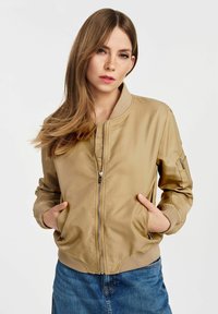 Beżowa kurtka bomberka wykonana z gładkiego materiału, z zamkiem błyskawicznym z przodu, ściągaczami przy mankietach, bocznymi kieszeniami oraz luźnym krojem.