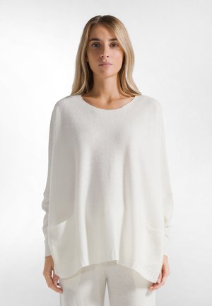 Pull oversized blanc en tissu tricot doux, avec un col large, des manches longues et deux poches latérales. Texture lisse, design minimaliste.