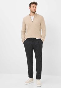 Beiger Pullover mit Reißverschluss am Hals aus weichem Stoff, kombiniert mit dunkelgrauen, maßgeschneiderten Hosen und hellen Sneakern. Ein einfaches, sauberes Design mit dezenten Strukturen.