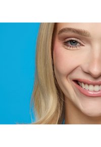 Nærbillede af en smilende kvinde med lyst blondt hår og blå øjne mod en klarblå baggrund.