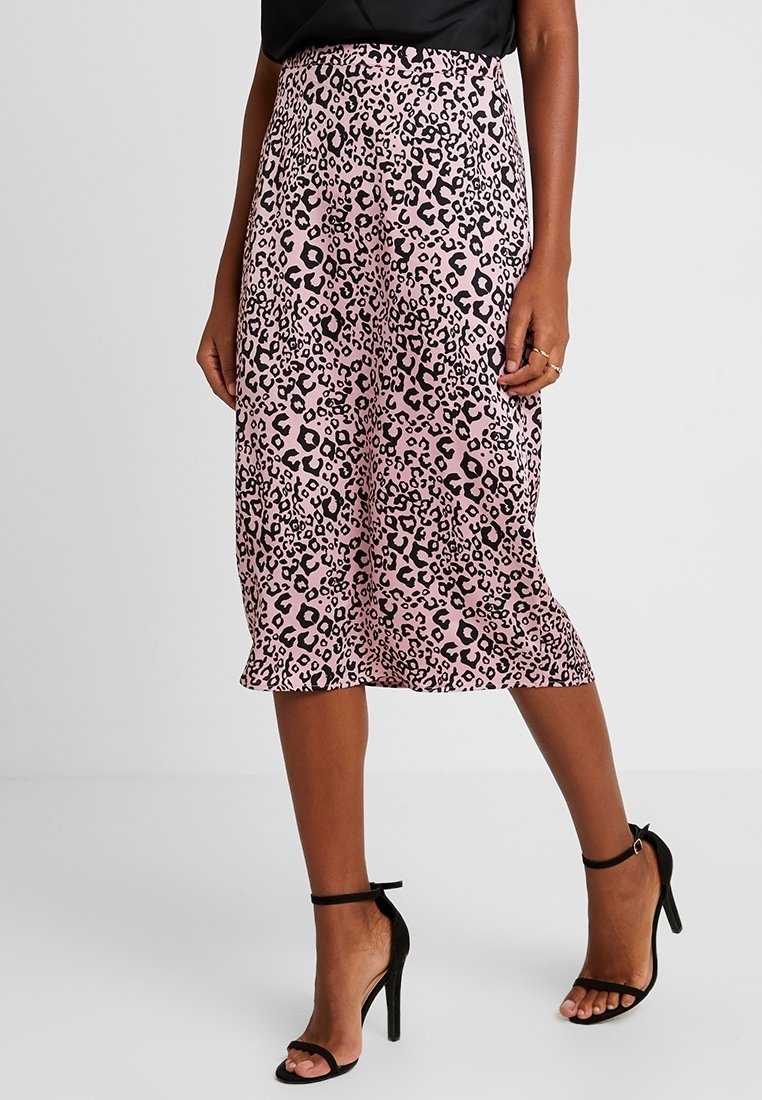 Femme portant une jupe midi rose avec imprimé léopard noir et des talons hauts noirs à bride à la cheville, debout devant un fond uni.