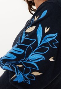 Pull marine avec broderies détaillées de motifs floraux bleus et beiges le long de la manche. Texture douce avec une coupe ajustée.