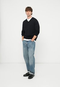 Crni džemper s V-izrezom preko bijele majice, uparen s lagano plavim širokim trapericama. Model nosi crne cipele, stoji na bijeloj pozadini.