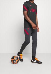 Czarny sportowy jersey z czerwonymi akcentami i logo, w połączeniu z ciemnoszarymi spodniami z czerwonym wykończeniem. Czarna i pomarańczowa piłka nożna przy stopie.