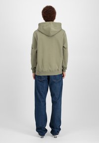Felpa verde oliva con cappuccio regolabile, polsini a costine e tasca laterale, abbinata a jeans blu larghi e sneakers.