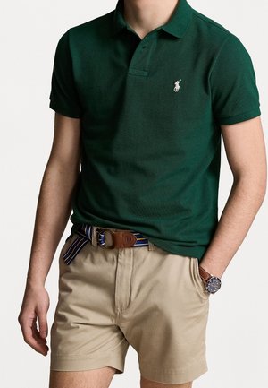 Mann trägt ein dunkelgrünes kurzärmeliges Polohemd, beige Shorts mit gestreiftem Gürtel und eine Armbanduhr, steht mit einer Hand in der Tasche.