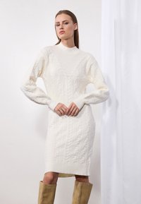 Robe en tricot torsadé couleur crème, avec un col haut, des manches longues et des poignets côtelés. Présente une silhouette ajustée et des motifs texturés sur l'ensemble.
