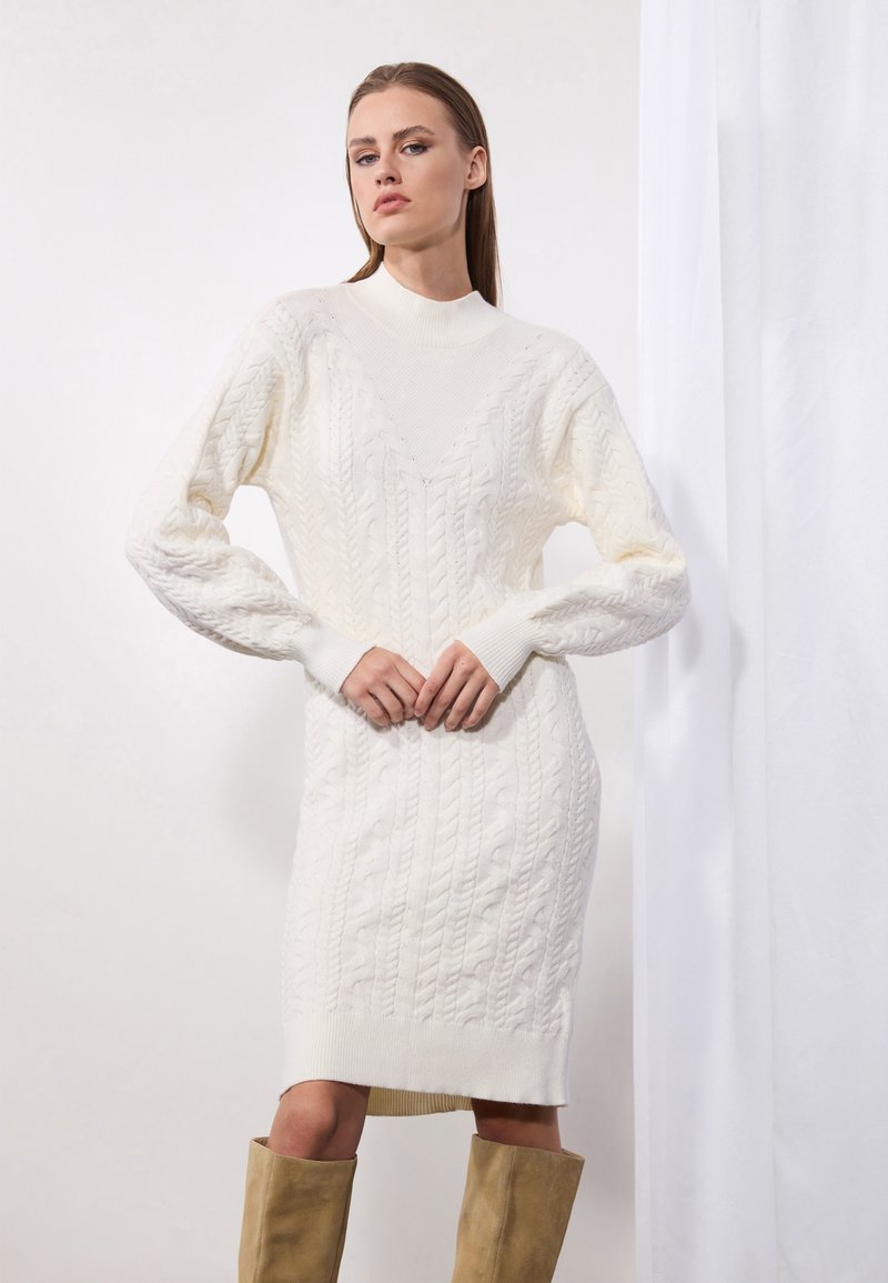 Robe en tricot torsadé couleur crème, avec un col haut, des manches longues et des poignets côtelés. Présente une silhouette ajustée et des motifs texturés sur l'ensemble.