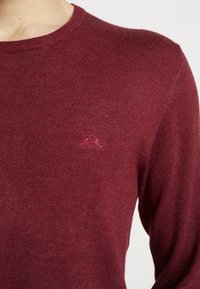 Viininpunainen neulepaita pyöreällä kaula-aukolla, jossa on pieni kirjailtu vuoristologo rinnassa ja pehmeä, teksturoitu pinta.