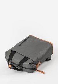 Grijze rugzak met een gladde afwerking, voorzien van zwarte banden, een tan leren accent en een ritsvak aan de zijkant voor extra functionaliteit.