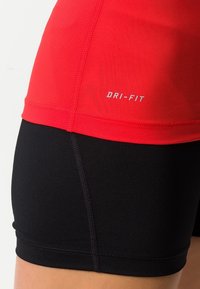 Punainen, hengittävä Dri-FIT-paita, joka on yhdistetty mustiin tiukasti istuviin shortseihin. Siinä on sileä kangas, kontrastivärit ja näkyvä brändin kirjotus.