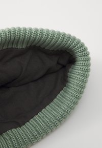 Bonnet en tricot avec un extérieur vert clair et une doublure en tissu foncé. Présente un bord côtelé pour un ajustement serré. Le matériau principal semble doux et texturé.
