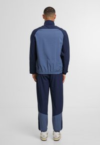 Jūras spēku sporta kostīms ar gaiši ziliem akcentiem, ietverzipažaketu, elastīgu jostasvietu un sašaurinātus bikses. Gluda auduma un brīva piegriezuma.