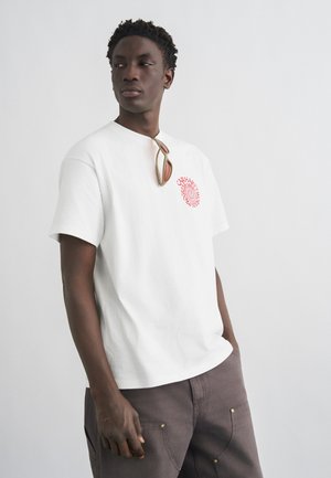 Mann trägt weißes T-Shirt mit rotem rundem Logo und Sonnenbrille, die am Kragen hängt, steht vor einfachem hellem Hintergrund.