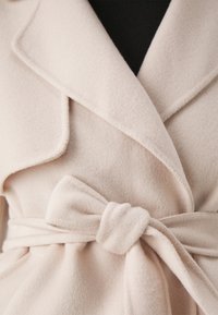 Vaaleanpunainen villakangastakki, jossa kietaisumallinen muotoilu, lovettu kaulus ja vyötäröllä vyö, jossa rusetti. Pehmeä pinta ja näkyvät ompeleet.