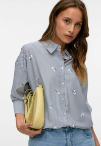 Camicia a righe blu e bianca con accenti floreali, abbinata a una borsa gialla. Il tessuto sembra leggero e strutturato.