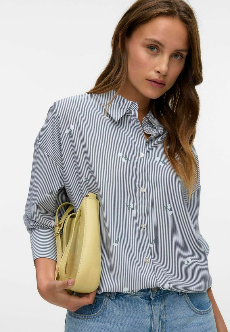 Camicia a righe blu e bianca con accenti floreali, abbinata a una borsa gialla. Il tessuto sembra leggero e strutturato.