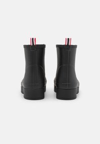 Schwarze Gummistiefel bis zum Knöchel mit einem klobigen Absatz, ausgestattet mit einer strukturierten Oberfläche und kontrastierenden ziehbaren Laschen in Rot, Weiß und Schwarz.