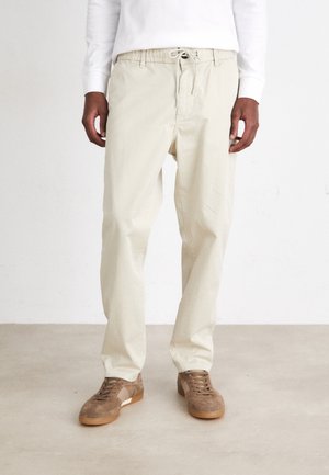 Pantalones en beige de corte relajado con cinturilla elástica y cordón, combinados con zapatillas de ante marrón claro que presentan un acento blanco.