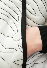 Gesteppte Jacke in Weiß mit wellenförmiger schwarzer Steppung. Verfügt über einen schwarzen Besatz und eine weiße Logo-Knopf nahe der Tasche. Weiche Stoffstruktur.