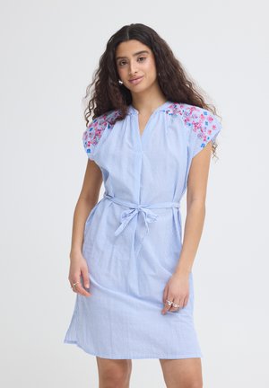 Donna con capelli lunghi e ricci che indossa un vestito a righe azzurro chiaro con ricamo floreale rosa e blu sulle spalle e una cintura da annodare in vita.