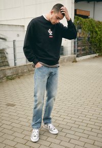 Sudadera negra con un gráfico y logo rojo, jeans de mezclilla azul claros y zapatillas a rayas negras y blancas sobre pavimento de adoquines.