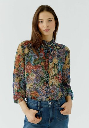 Jeune femme aux longs cheveux bruns portant un chemisier floral multicolore et un jean bleu, debout les mains dans les poches devant un fond uni.