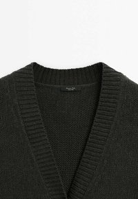 Tummanvihreä neulottu neuletakki, jossa syvä V-pääntie ja resorireunus. Tekstuuri on pehmeä ja paksu, ja se esittelee monimutkaisen kudontakuvion.