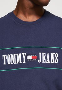 T-shirt in cotone blu navy con cuciture accentate verdi, che presenta un grande testo ricamato bianco "TOMMY JEANS" sopra un piccolo patch con logo.