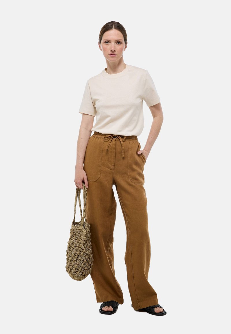 T-shirt beige à manches courtes associé à un pantalon brun ample avec taille à cordon. Le modèle tient un sac en osier texturé.