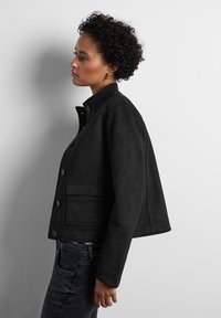 Svart mockajacka med hög krage, knäppning framtill och en stor ficka. Designen är kort och skräddarsydd, vilket framhäver en slät textur.