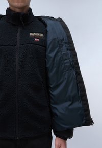 Chaqueta de forro polar negra con textura estructurada, que presenta un cierre con cremallera, forro interior en color marino y logo bordado en el lado izquierdo del pecho.
