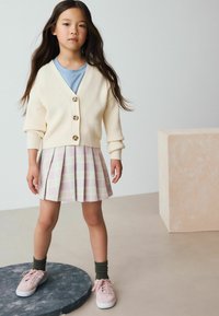 Cardigan en ivoire à côtes avec des boutons, t-shirt bleu clair, jupe plissée à carreaux rose, et baskets roses avec chaussettes vertes. La tenue présente des textures superposées.